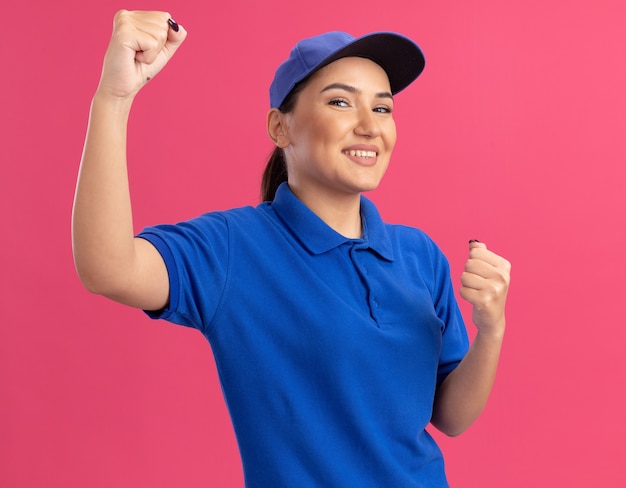 Feliz y emocionada joven mujer de entrega en uniforme azul y gorra apretando los puños de pie sobre la pared rosa