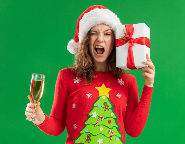 Feliz y emocionada joven en rojo suéter de Navidad y gorro de Papá Noel sosteniendo una copa de champán y presente de pie sobre fondo verde