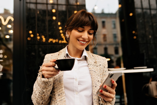 Feliz emocionada joven vestida de chaqueta blanca está usando teléfonos inteligentes y auriculares mientras está tomando café en el fondo de las luces de la ciudad Foto de alta calidad