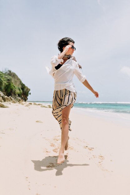 Feliz emocionada joven vestida con pantalones a rayas, camisa blanca y gafas saltando en la playa con arena blanca cerca del océano