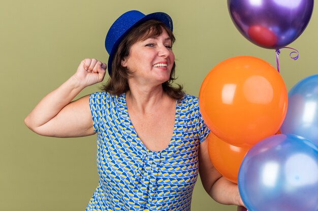 Feliz y emocionada mujer de mediana edad con sombrero de fiesta con un montón de globos de colores divirtiéndose sonriendo alegremente celebrando la fiesta de cumpleaños de pie sobre la pared verde