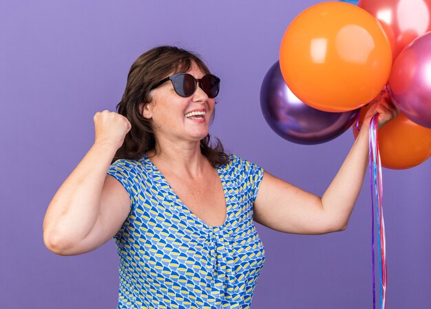 Feliz y emocionada mujer de mediana edad sosteniendo un montón de globos de colores apretando el puño celebrando la fiesta de cumpleaños de pie sobre la pared púrpura