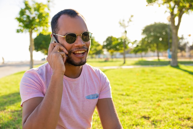 Feliz emocionado chico latino en gafas de sol hablando por teléfono móvil