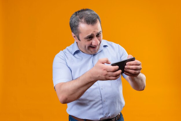 Feliz y emocionado hombre de mediana edad en camisa azul a rayas verticales jugando en su teléfono móvil mientras está de pie