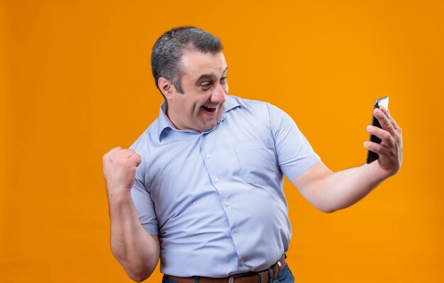 Feliz y emocionado hombre de mediana edad vestido con camisa azul a rayas verticales mirando su teléfono móvil y levantando la mano en el gesto de puño cerrado mientras está de pie sobre una espalda naranja