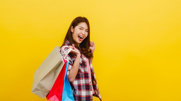 Feliz emocionado joven asiática llevando bolsas de compras con la mano levantando en ropa casual y mirando a la cámara sobre la pared amarilla. Concepto de expresión facial, venta estacional y consumismo.