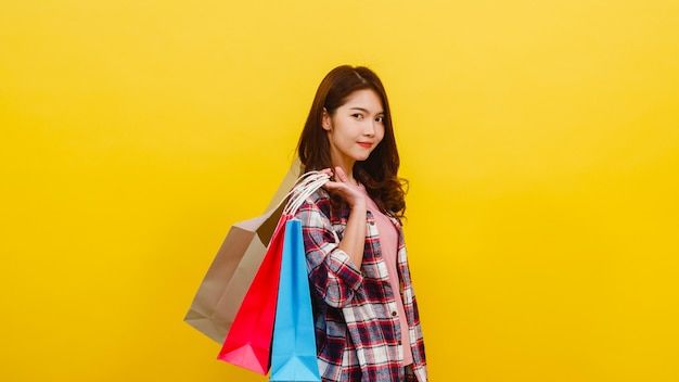 Feliz emocionado joven asiática llevando bolsas de compras con la mano levantando en ropa casual y mirando a la cámara sobre la pared amarilla. Concepto de expresión facial, venta estacional y consumismo.