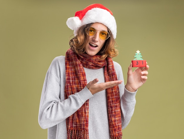 Feliz y emocionado joven vestido con gorro de Papá Noel de Navidad y gafas amarillas con cálida bufanda alrededor de su cuello presentando cubos de juguete con fecha de año nuevo de pie sobre pared verde