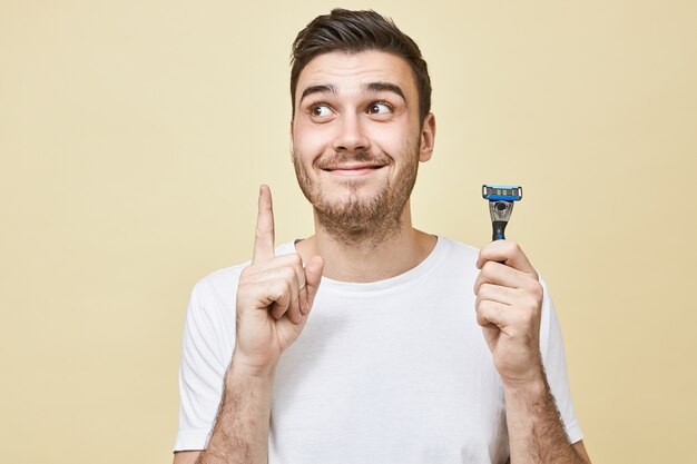 Feliz emotioanl joven con barba posando vistiendo una camiseta blanca sosteniendo el dedo levantado como si tuviera una buena idea mientras se afeita la cara en el baño, usando una maquinilla de afeitar, mirando hacia arriba y sonriendo