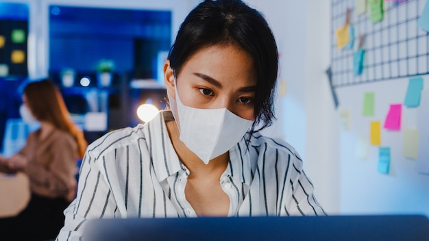 Feliz empresaria de Asia con mascarilla médica para el distanciamiento social en una nueva situación normal para la prevención de virus mientras usa la computadora portátil en el trabajo en la noche de la oficina.