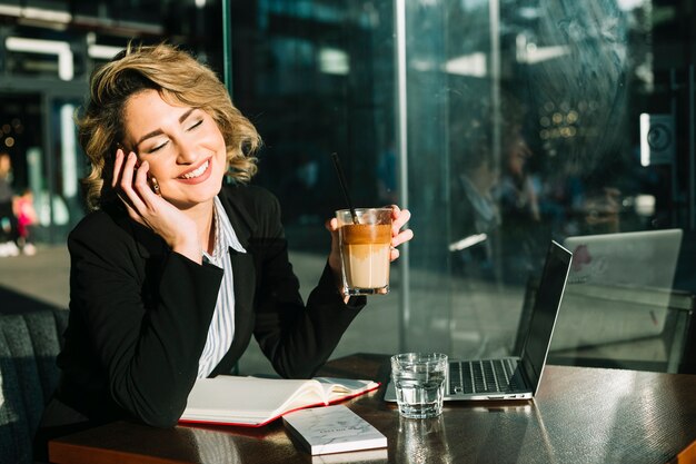 Feliz empresaria hablando por teléfono inteligente mientras sostiene un vaso de batido de chocolate en un restaurante