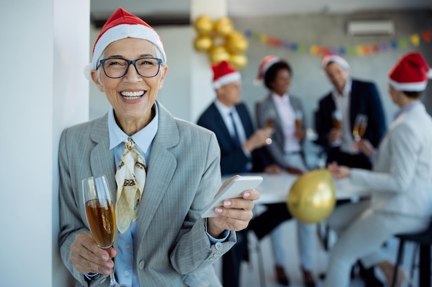 Feliz empresaria madura usando teléfono móvil en la fiesta de Año Nuevo en la oficina