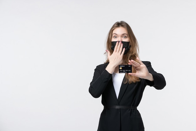 Feliz empresaria en traje con su máscara médica y mostrando la tarjeta bancaria haciendo gesto de silencio en blanco aislado