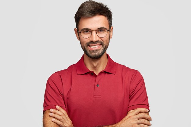 Feliz empresario masculino confiado con una sonrisa positiva, tiene barba y bigote, mantiene los brazos cruzados, está de buen humor después de una reunión exitosa con sus socios, posa contra la pared blanca, vestido de manera informal