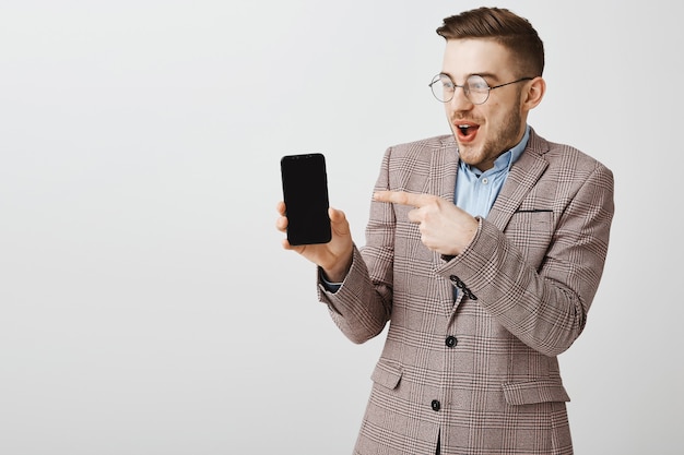 Feliz empresario masculino en traje apuntando con el dedo a la pantalla del teléfono inteligente, mostrando la aplicación móvil