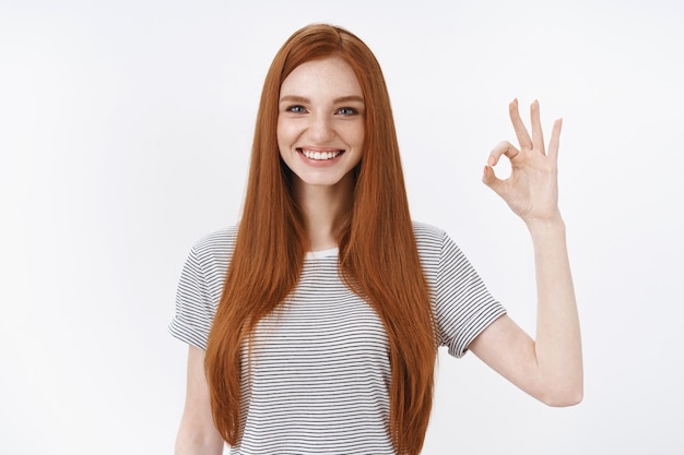 Feliz, encantadora, atractiva pelirroja estudiante da una respuesta positiva, muestra bien, bien, gesto normal, expresa una buena actitud positiva, como resultado, pared blanca de pie