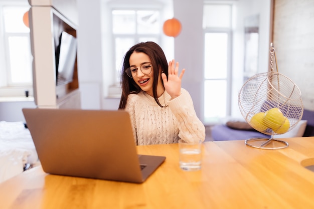 Feliz encantadora joven está sentada en la cocina y trabajando en su computadora portátil y teléfono móvil