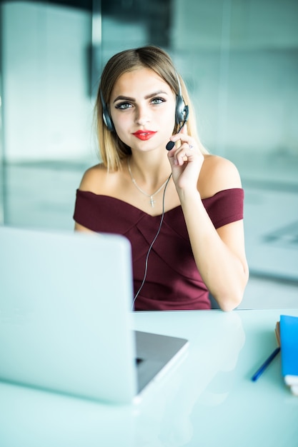 Feliz encantadora joven sentada y trabajando con un portátil con auriculares en la oficina