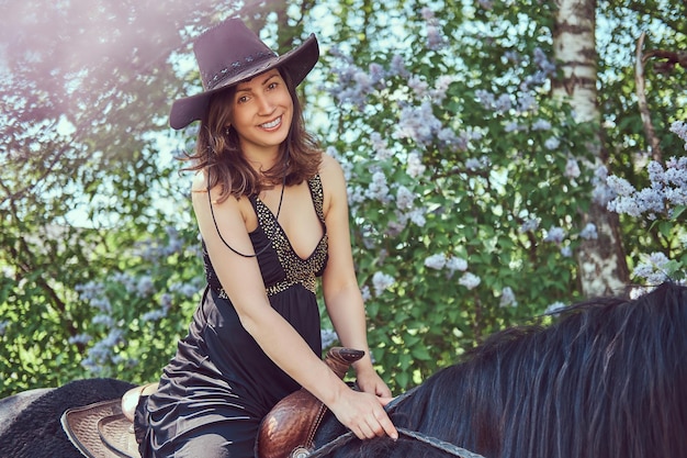 Feliz y encantadora morena con ropa negra y sombrero montando un caballo marrón en el jardín de flores.