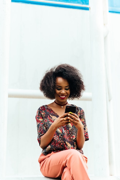 Feliz encantadora mujer afroamericana, mirando la pantalla del teléfono inteligente, sonriendo alegremente