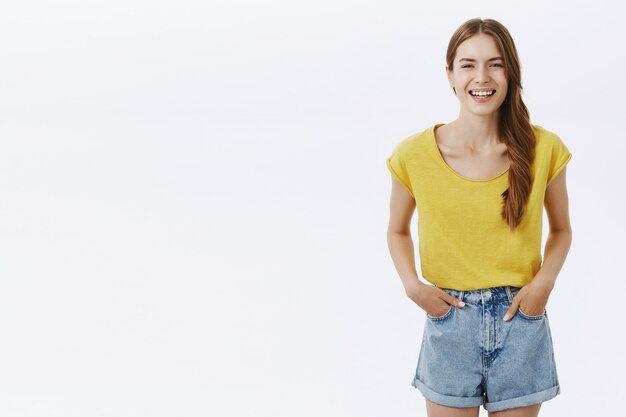 Feliz encantadora mujer de aspecto amistoso con el pelo trenzado posando en camiseta amarilla y pantalones cortos sonriendo encantado tomados de la mano en los bolsillos de pie casualmente