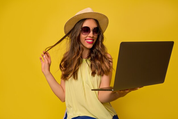 Feliz encantadora mujer europea con sombrero y gafas de verano trabajando con ordenador portátil