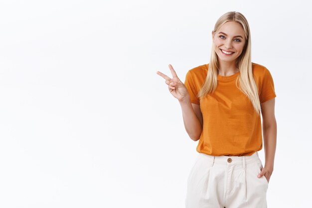 Feliz, encantadora mujer rubia con estilo femenino en pantalones de moda de camiseta naranja, tome la mano en el bolsillo, muestre la paz o el signo de la victoria como una sonrisa modesta con una expresión encantadora y satisfecha, soporte fondo blanco