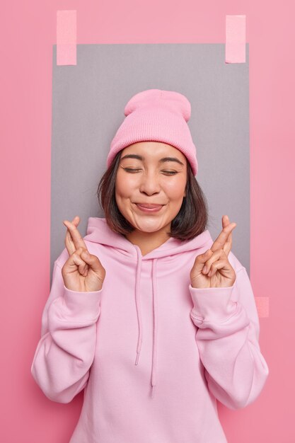 Feliz y esperanzada joven asiática mantiene los dedos cruzados cree en la buena suerte anticipa algunos resultados cierra los ojos vestidos con una cómoda sudadera con capucha rosa y poses de sombrero contra la pared vacía en blanco