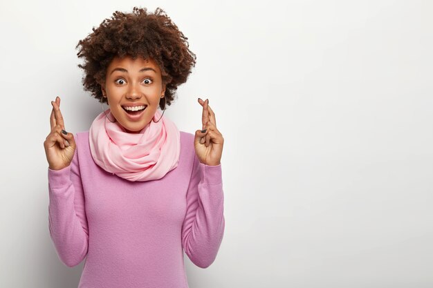 Feliz y esperanzada mujer afro mantiene los dedos cruzados, desea tener suerte en la entrevista de trabajo, viste cuello alto púrpura y pañuelo de seda