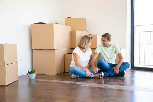 Feliz esposa y esposo sentados con las piernas cruzadas en el piso en un apartamento nuevo cerca de cajas de cartón