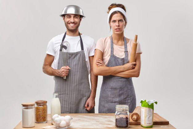 Feliz esposo en delantal sostiene un batidor, la esposa enojada mantiene las manos cruzadas, se para cerca de la mesa de la cocina, preparan la cena juntos, rodeados de todos los ingredientes necesarios, aislados en la pared blanca