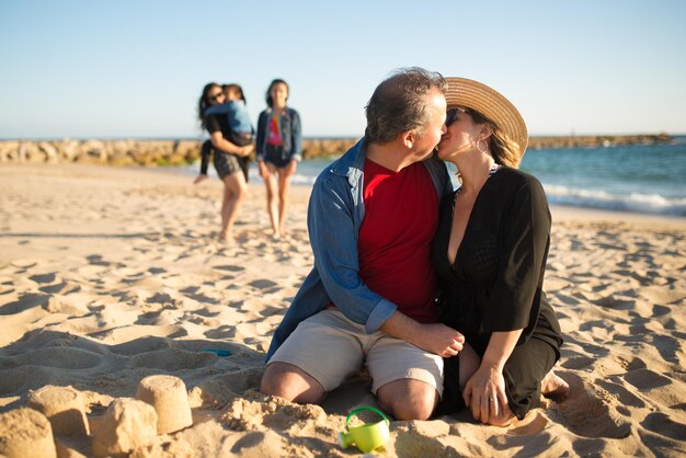 Feliz esposo y esposa besos en la playa. Hombre y mujer, sentar arena, abrazar