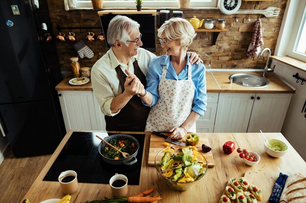 Feliz esposo y esposa mayores comunicándose mientras se toman de la mano y preparan una comida en la cocina