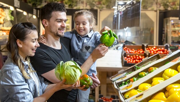 Feliz esposo y esposa con un niño compran verduras. Familia alegre de tres eligiendo pimiento y verduras en el departamento de verduras del supermercado o mercado.