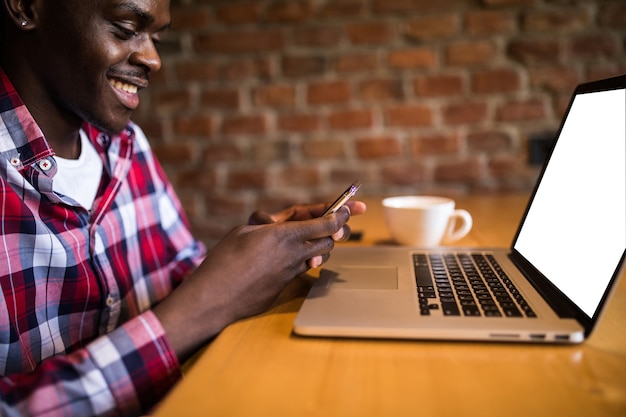 Feliz estudiante universitario afroamericano con linda sonrisa escribiendo un mensaje de texto en un dispositivo electrónico, sentado en el café tablein cafe.