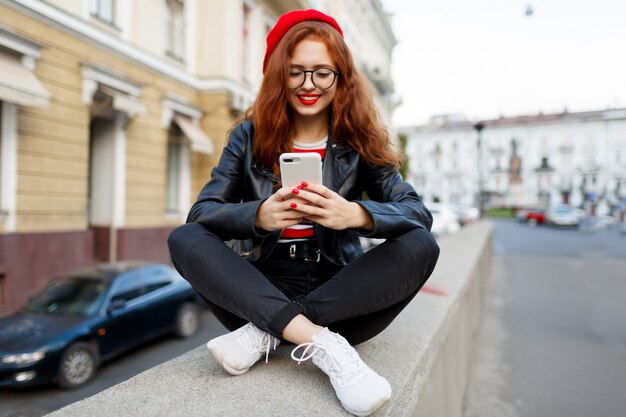 Feliz fabulosa mujer de jengibre en elegante boina roja en la calle con smartphone