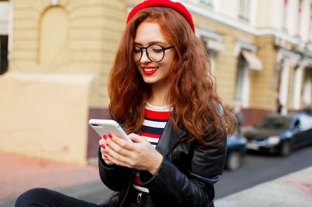 Feliz fabulosa mujer de jengibre en elegante boina roja en la calle con smartphone