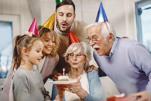 Feliz familia extendida soplando velas en el pastel de cumpleaños mientras celebra una fiesta de cumpleaños en casa