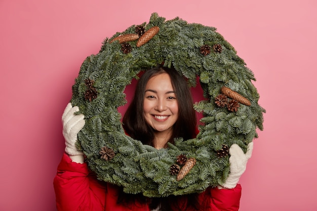 Feliz floristería asiática realiza una clase magistral sobre cómo hacer decoraciones navideñas para el hogar, mira con alegría a través de una corona de piel hecha a mano