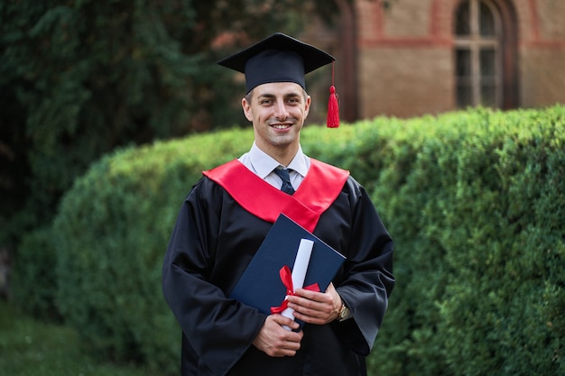Feliz graduado masculino caucásico en graduación resplandor con diploma mirando a cámara en el campus.