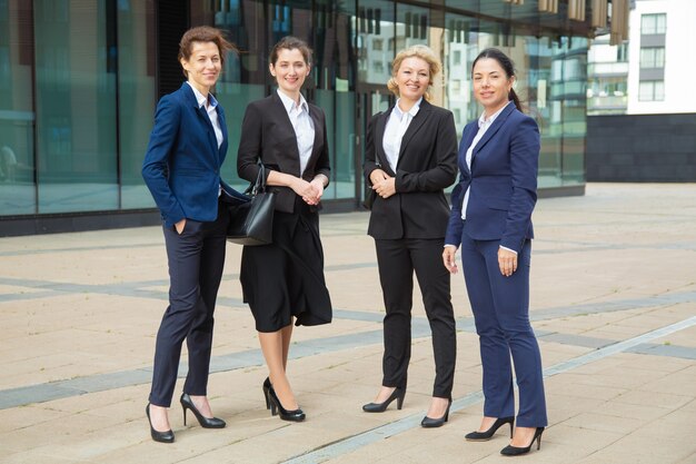 Feliz grupo empresarial femenino exitoso de pie junto al edificio de oficinas, posando, mirando a cámara y sonriendo. Vista frontal de cuerpo entero. Concepto de retrato de grupo de mujeres empresarias