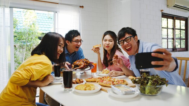 Feliz grupo de jóvenes almorzando en casa. Fiesta familiar de Asia comiendo pizza y haciendo selfie con sus amigos en la fiesta de cumpleaños en la mesa de comedor juntos en casa. Celebración de vacaciones y unión.