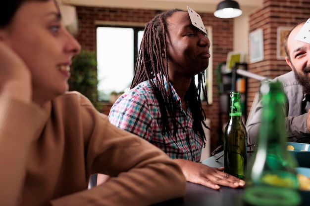 Feliz grupo multicultural de amigos sentados en casa mientras juegan a adivinar juegos de cartas juntos. jugadores adultos jóvenes que disfrutan de actividades de ocio divertidas mientras toman bocadillos y bebidas.