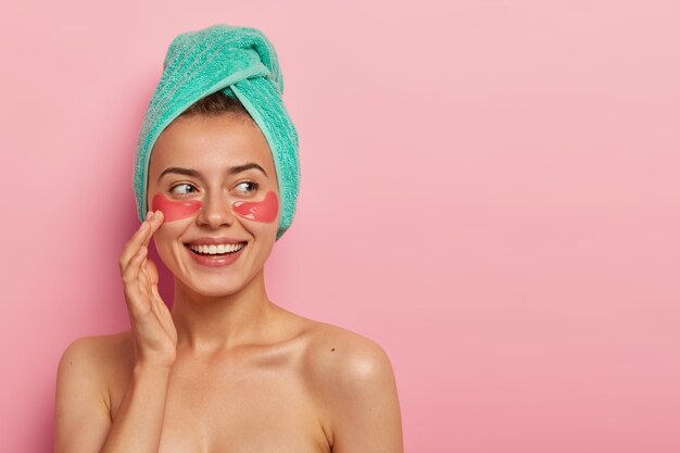 Feliz y guapa mujer joven toca suavemente la cara, sonríe agradablemente, dedica su tiempo libre durante el fin de semana a tratamientos de belleza, hidrata la delicada piel debajo de los ojos, se preocupa por el cuerpo y la tez