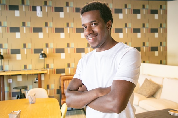 Feliz guapo afroamericano posando con los brazos cruzados en el interior de la cafetería o el coworking, mirando a la cámara y sonriendo