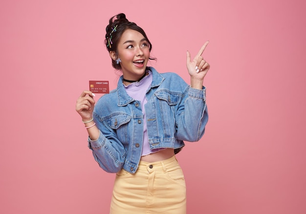 Feliz hermosa adolescente asiática adicta a las compras mostrando tarjeta de crédito y su dedo apuntando en rosa