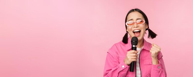 Feliz hermosa chica asiática cantando con micrófono usando micrófono disfrutando de karaoke posando sobre fondo de estudio rosa