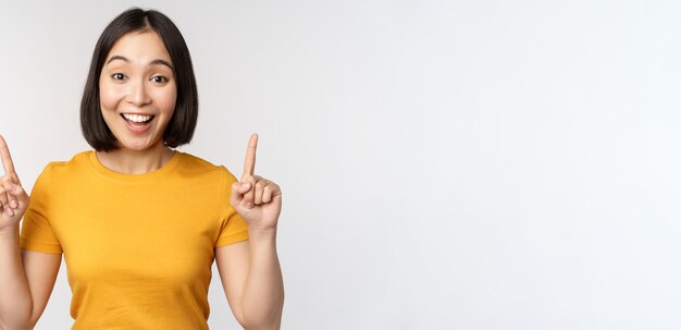 Feliz hermosa chica asiática mostrando publicidad apuntando con el dedo hacia arriba de pie sobre fondo blanco.