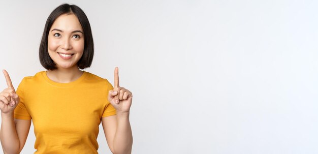 Feliz hermosa chica asiática mostrando publicidad apuntando con el dedo hacia arriba de pie sobre fondo blanco.