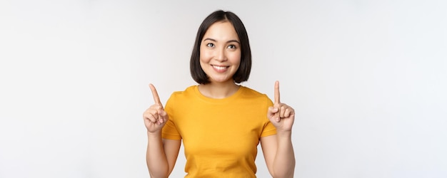 Feliz hermosa chica asiática mostrando publicidad apuntando con el dedo hacia arriba de pie sobre fondo blanco.